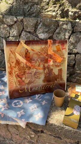 The video shows a picnic setup against a stone wall. A tote bag with a "Montserrat" design rests on a blue patterned cloth. Next to it is a small paper cup. The camera pans right to reveal an arrangement of items for the picnic. These include a bottle of "Abadia de Biowmont" sparkling wine, two boxes of "1928 Fatget" salchichón with truffle, a package of bread sticks, a sausage, and some crackers. The background is a rough stone wall.