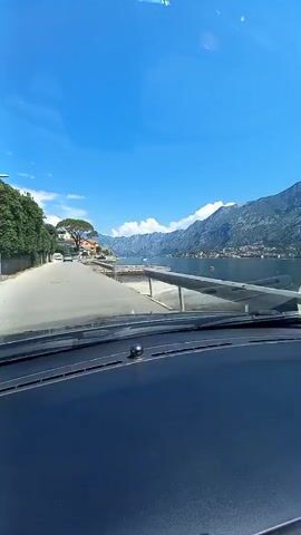 The video captures a view from inside a vehicle looking out the windshield. The vehicle appears to be driving on a paved road that runs alongside a body of water, likely a bay or a large lake, with mountains in the background. The road curves gently to the left as the vehicle moves forward. On the left side of the road, there are buildings and lush green trees. On the right side, the water stretches out, with several boats docked or moored. The water reflects the bright blue sky, which is dotted with a few white clouds. The mountains in the distance are large and rugged, with some structures visible on their slopes. The sun is shining brightly, creating reflections on the windshield and in the water. The overall impression is a scenic drive through a coastal or lakeside area on a clear day.