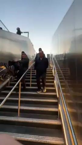 The video shows several people walking down a long outdoor staircase. The stairs have metal handrails, and the walls on either side are reflective, adding to the brightness of the scene. The sky is visible above, appearing bright and clear, suggesting it is daytime. The people are dressed casually, wearing jackets and trousers. They are walking at a normal pace, with some looking towards the camera while others are focused on the stairs. The lighting is good, and the video is stable, providing a clear view of the people and the environment.