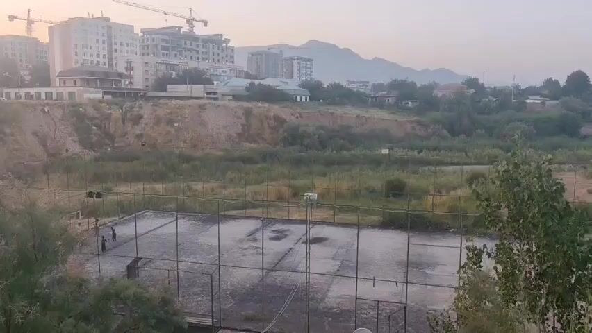 The video shows an outdoor sports court, likely a basketball or tennis court, with a chain-link fence surrounding it. The court surface appears to be asphalt and has some wet patches and debris on it. There are two figures, appearing to be children, on the court, possibly playing. Beyond the court, there is a patch of overgrown vegetation, followed by a dirt embankment. In the background, several multi-story buildings and some smaller houses are visible, with mountains forming the backdrop under a hazy sky, suggesting either early morning or late afternoon lighting. A construction crane is visible near one of the buildings. The overall impression is of an urban or semi-urban environment with some neglect or disuse.