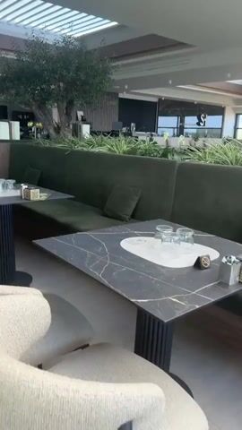 The video pans across a dimly lit, modern restaurant interior. In the foreground, two plush cream-colored chairs with curved backs are visible. A marble-topped table with a black pedestal base stands between them. Behind this, a long, dark green velvet banquette seating area curves around the room, interrupted by a large artificial tree. The banquette has dark marble-topped tables. In the background, a bar area is visible with a bartender, and other patrons. The overall ambiance is sophisticated and contemporary.