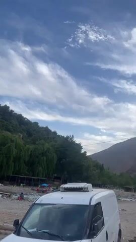 The video shows a landscape with a blue sky dotted with scattered clouds. A rocky mountain rises in the background, covered with green vegetation. A white delivery van with a cooling unit on top is parked in the foreground. In the middle ground, there are small buildings and trees, with people visible in the distance. As the video progresses, it pans to reveal more buildings nestled on the mountainside. The overall environment appears to be a rural or mountainous area. The camera movement is smooth and deliberate, focusing on the natural scenery and the human presence within it.