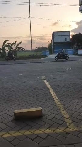 The video shows a street scene at dusk. In the foreground, there is a paved road with interlocking brick pavers and yellow road markings. A rectangular object, possibly a curb or a marker, is placed on the pavers. In the middle ground, two motorcycles are visible. The one on the left carries two people, while the one on the right appears to have a single rider. Beyond the road, there is a field with some vegetation, possibly rice paddies, and banana trees. A building with a blue facade is also present on the right side of the frame. A tall lamppost stands near the center of the scene. The sky is a soft orange and pink, indicating sunset. The overall atmosphere is calm and rural.