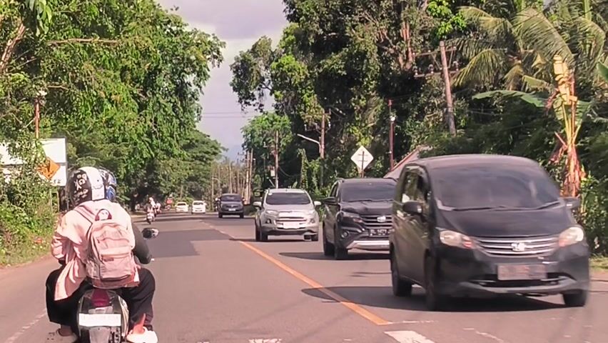 The video captures a daytime scene from the perspective of a vehicle, likely a motorcycle, traveling along a two-lane asphalt road in Reuhattuha, Indonesia. The road features a solid yellow line separating the directions of traffic. Lush green tropical trees and foliage densely line both sides of the road, with utility poles and overhead wires also visible.

In the immediate foreground, the back of a person riding a motorcycle is visible. They are wearing a light-colored top, a backpack, and a helmet adorned with a cartoonish design. The video primarily focuses on oncoming traffic.

At the start, a dark-colored SUV, possibly a Daihatsu Terios or Toyota Rush, passes on the opposite side of the road. It is followed by a silver Ford EcoSport SUV. As the camera's perspective moves forward, the silver SUV passes, and then a gray Toyota Kijang Innova minivan approaches and drives past. In the distance, other vehicles, including cars and motorcycles, can be seen on the road.

Notable details include a white diamond-shaped road sign on the right side of the road, displaying a black symbol resembling a railroad crossing warning (an 'H' crossed by two horizontal lines). At approximately 0:02, a small, white rectangular sign with the text "BBM" in black letters is visible on the left side of the road, situated near a small structure or shack. The weather appears clear or partly cloudy, indicating ample daylight. No specific emotions are discernible from the drivers or riders.