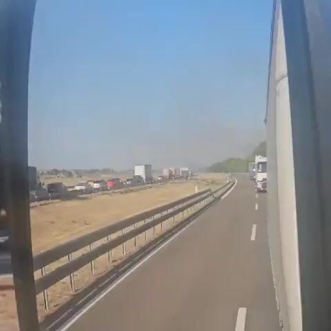 The video shows a rearview mirror reflection of a highway with heavy traffic congestion. A long line of cars and trucks are stopped in traffic. Smoke or dust can be seen in the distance, possibly indicating a fire or other event causing the slowdown. The sky is clear and blue, suggesting it is daytime. The camera appears to be mounted on a vehicle, likely a truck, given the perspective. The road is paved and has guardrails. Vegetation is visible on the side of the road. The scene suggests a disruption on the highway leading to a standstill.