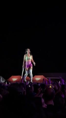 A female performer is on a stage in front of a dark background. She is wearing a light green crop top, pink shorts, and white thigh-high socks. She has dark hair pulled into two pigtails. She is holding a microphone in her right hand and what appears to be a prop resembling a baseball bat with her left. She is looking towards the left side of the frame and appears to be singing or speaking. In the foreground, a crowd of people are visible, many with their hands raised, holding up cell phones to record. In the background, behind the performer, are orange structures that appear to be part of the stage setup. The lighting is dim, with spotlights focused on the performer.
