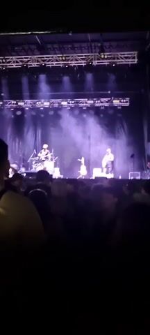 A concert crowd is visible in the foreground, facing a stage where a band is performing. Bright stage lights, beams of white and purple, illuminate the band members and create a haze. The crowd appears to be enjoying the performance, with some individuals dancing. The overall atmosphere is energetic and lively.