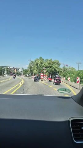 The video appears to be taken from inside a vehicle, looking forward and to the side. Several motorcycles are visible on a road with yellow lane markings and construction barriers. The sky is clear and blue, with green trees and buildings in the background. There is also a construction crane visible. The motorcycles are being ridden by people wearing helmets and some appear to be part of a group or procession.