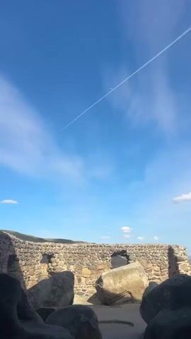 The video shows a clear, bright blue sky with wispy clouds. A contrail cuts across the sky at an angle, running from the lower left to the upper right. The moon, a thin crescent, is visible slightly above and to the right of the contrail. At the bottom of the frame, a low, sandy hill is visible. There are also some small puffy clouds in the lower right corner. The camera appears to be held and angled upwards, capturing the sky and horizon.