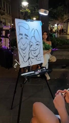This video captures an artist drawing a caricature of a couple on a street at night. The artist, a woman with blonde hair and a large octopus tattoo on her arm, is actively drawing on a white canvas propped on a tripod easel. A woman in a green top sits behind the easel. The drawing itself depicts a man and a woman with exaggerated features, typical of caricatures. The background shows a bustling street scene with other people and lights, indicating an outdoor urban environment, possibly a pedestrian zone or promenade. There are drawing supplies, including markers, on a tray attached to the easel. The scene is illuminated by artificial lights, including a spotlight directed at the canvas and streetlights.