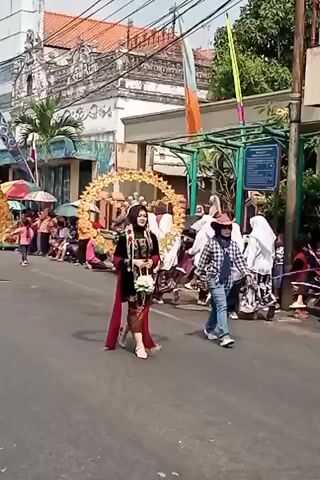 A parade is taking place on a paved street, lined with buildings and spectators. In the foreground, a woman wearing a black hijab, a maroon dress with gold trim, and a circular gold headdress adorned with sunflowers walks down the street. She is holding a bouquet of white flowers. To her right, a person in a plaid shirt, blue jeans, and a wide-brimmed hat walks in the same direction. In the background, other participants in traditional attire, some wearing hijabs, walk in the parade, and spectators are seated on the side of the street, some holding umbrellas. Colorful flags and banners are visible along the street. The buildings appear to be old, with ornate architectural details. The sky is bright and sunny.