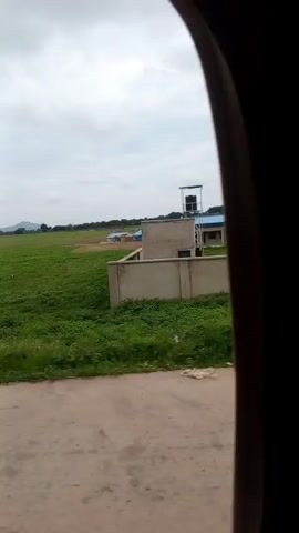 The video shows a wide, green, grassy field under a cloudy sky. In the foreground, there is a concrete road or path. In the distance, there are some trees and a few buildings, possibly houses or structures under construction. A tall, solitary tree stands in the middle of the field. A concrete wall or fence runs along the right side of the field. The video appears to be taken from a moving vehicle.