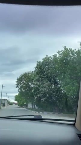 The video shows a drive down a narrow paved road in a rural area. The view is primarily through the car's windshield, showing the road ahead and the passing scenery. Houses with brick facades and pitched roofs line the road, interspersed with trees. The sky transitions from overcast to partly cloudy with patches of blue. There are utility poles with wires running along the road. The car's dashboard is visible, with a rearview mirror and an air freshener hanging from it. There's music playing in the background.