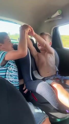Two young boys are in the back seat of a car. The boy on the left is wearing a striped shirt and jeans. The boy on the right is shirtless and wearing shorts. They are holding hands and appear to be playing or interacting playfully. The car interior is visible, with a car seat in the background. Sunlight streams in from the windows, suggesting it is daytime.