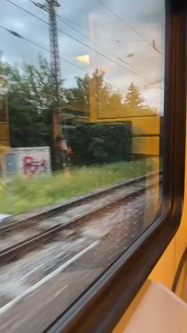 The video shows a view from a train window as it travels. The foreground is dominated by the train tracks, which appear blurred due to the motion. Beyond the tracks, there is a grassy area with some bushes and trees. In the background, a building with the text "KULTURFABRIK" is visible. The sky is overcast, suggesting it might be a cloudy day. The overall impression is a peaceful journey through a rural or semi-rural landscape. There are no people clearly visible in the frame. The light suggests it is daytime, possibly late afternoon or early evening, given the soft lighting.