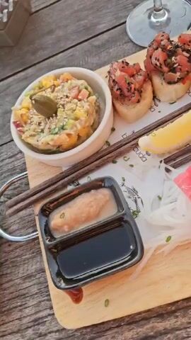 The video shows a wooden board with an assortment of food and drinks on it, set on a wooden table. In the top left, there is a white bowl filled with a salad-like mixture. Next to it are four pieces of bread topped with diced tuna and black sesame seeds. Two lemon wedges rest on the board in between the bread and the tuna. Below that, there is a small, black divided dish with a light brown sauce in one section and dark sauce in the other. Fresh tuna sashimi slices are neatly arranged on a bed of shredded white radish. A small flower-shaped carrot with wasabi in the middle sits atop the tuna. To the right of the sashimi is a small metal basket filled with fried tortilla chips. Behind the board, there are two drinks: one in a wine glass, possibly an aperol spritz, and another in a highball glass with a straw and a slice of watermelon. The table is made of weathered wood. Sunlight is visible in the background.