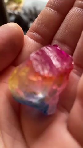 A person holds a colorful, irregular chunk of crystal in their palm. The crystal features distinct bands of blue, yellow, pink, and red hues, suggesting an artificial coloring process. The lighting accentuates the gem's translucent quality and uneven surface. The person's fingers grip the crystal, providing a scale for its size. The background is blurred, revealing only indistinct shapes.