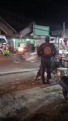 A man wearing a dark jacket with a circular emblem on the back stands facing away from the camera, near a motorcycle. He is on the side of a road at night. Across the road, there are several vehicles, including a white van and what appears to be a food stall with people around it. A motorcycle is seen passing by in the middle ground. The scene is lit by artificial lights, suggesting it is an urban or semi-urban area.