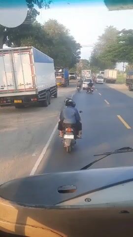 The video shows a rear view of a motorcycle with two occupants, a driver and a passenger, riding in the middle of a two-lane road. The motorcycle is traveling in the same direction as a line of vehicles, including trucks and cars. The driver is wearing a blue shirt and a helmet, and the passenger is also wearing a helmet. The road appears to be paved and is lined with trees on either side. The sky is visible in the background and appears to be clear and sunny. The overall impression is of traffic congestion.