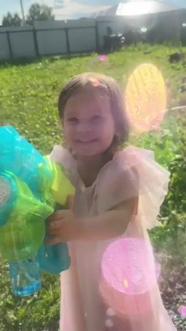 A young girl in a pink and white dress smiles at the camera while holding a blue and green bubble gun. Bubbles are floating around her. The girl is standing in a grassy yard with a fence in the background. The sun is shining.