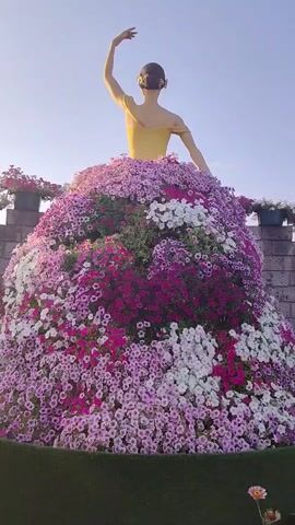 A statue of a ballerina in a yellow leotard stands with her arms outstretched, her body covered in a voluminous skirt made of pink and white flowers. The statue is positioned outdoors, with a clear blue sky in the background and stone structures on either side. The flowers form a dense, rounded shape around the statue's lower body, creating the illusion of a flowing gown.