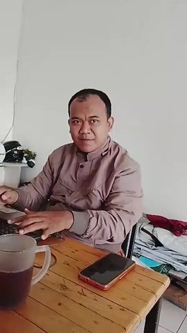 The video opens on a man in a light brown jacket seated at a wooden table, typing on a laptop. A mug of dark liquid sits to his left, and a smartphone is on the table to his right. In the background, a white wall is visible, with a shelf holding various items including a fan, a white object that appears to be a helmet, and a black bag. A calendar is also on the wall, displaying the year 2026.

The camera then pans to the left, revealing another man wearing a blue sweatshirt and a baseball cap. He is also seated at the table, holding a cigarette in his right hand and a mug in his left. A white power strip with multiple outlets is placed in front of him, with various cables connected. He appears to be looking down.

The camera continues to pan, focusing on the second man, who then brings the cigarette to his lips. The view shifts again, showing a closer shot of the second man with the cigarette in his mouth. A white calendar is now visible on the table in front of him, along with the power strip.

Finally, the camera pans to the right, showing a broader view of the table. An orange envelope is placed on top of some papers. A white calendar is also on the table. In the background, a black chair is visible against a white wall. The video ends with a view of the wooden table.
