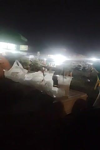 The video depicts an outdoor setting at night, possibly a market or public gathering, as evidenced by the presence of vendors and blurred figures in the background under bright lights. The camera focuses on a table with a black paper coffee cup with a lid and straw. There's also a clear plastic glass with a straw nearby. Keys are on the table next to the coffee cup. The foreground is dark, suggesting the perspective of someone sitting at the table, while the background remains out of focus due to the depth of field.