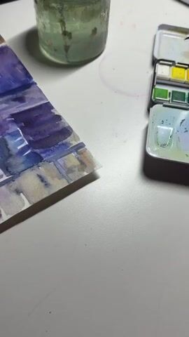 The video shows a close-up of a person's hand painting a watercolor picture. The hand, with bright yellow nail polish, is holding a paintbrush and applying paint to the paper. The painting appears to be a landscape with shades of purple, blue, and brown. Next to the painting is a watercolor paint palette and a jar of water. The scene is set on a white surface, likely a table. The lighting suggests an indoor environment.