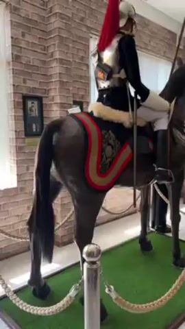 The video showcases a static display inside what seems to be a museum or historical site. The primary focus is a life-sized model of a horse with a mannequin dressed in a historical military uniform riding it. The horse is dark brown, complete with a decorative saddle blanket featuring red and gold trim. The mannequin rider wears a silver breastplate, a helmet with a red plume, a black jacket, white riding breeches, and tall black boots. A long spear is positioned upright beside the rider. The display is surrounded by a faux green grass mat and secured by a rope barrier. In the background, there is a brick wall with a window, and partially visible behind the horse and rider, there is another mannequin standing in a dark uniform. The overall impression is of a meticulously arranged exhibit meant to depict a historical military figure on horseback.