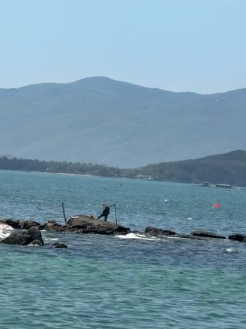 A long shot captures a person wearing a hat and backpack, standing on a dark, rugged rock in the calm blue sea. This rock is part of a larger formation, with other rocks visible to the left. The person appears to be engaged in an activity involving two wooden poles, one on either side of them, positioned within the water or attached to the rocks. In the background, a long, narrow strip of land, likely a beach, stretches across the horizon, lined with trees and buildings. Beyond this land, large, green mountains rise up against a clear, light blue sky. A red buoy and a few small boats are visible in the distance on the water. The sun is shining brightly, creating reflections on the water's surface.