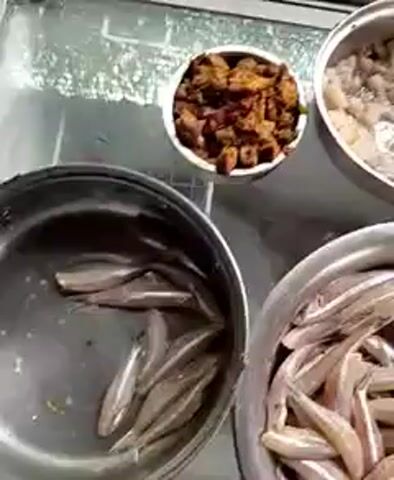 This video shows a close-up, overhead view of several stainless steel and blue bowls filled with various food items. The first bowl, at the top left, contains a pale, granular substance that appears to be cooked pasta or grains, possibly macaron. To its right, a large bowl is filled with what looks like small, whole fish, silvery in appearance with thin dark stripes. Below the pasta bowl, another bowl, this one blue, is packed with a similar pale, granular food item, also likely pasta. In the bottom center of the frame, a stainless steel bowl is filled with more small, whole fish, similar to those in the upper right bowl. The items are arranged on a reflective surface that appears to be a countertop or display area, possibly in a market or food stall. The lighting is dim, creating a slightly grainy effect on the footage. There are no people visible in the frame.