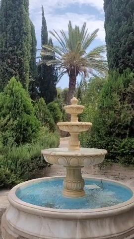 This is a video of a tiered fountain in a garden setting. The fountain is made of a light-colored stone and has three basins, with water flowing from the top basin down to the lowest one, which is filled with blue water. Surrounding the fountain are lush green plants, including tall cypress trees, a palm tree, and various bushes and smaller plants. The sky is visible in the background with some clouds. The video pans slowly to the left, showing more of the garden and its diverse greenery. There are no people visible in the video. The overall impression is one of a peaceful and well-maintained garden.