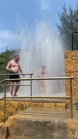 The video shows a middle-aged man and a young woman in swimwear standing in a large fountain. The man is heavier set and wearing black shorts and white shoes, while the woman is wearing a black bikini. They both appear to be enjoying the water, with the woman initially reacting with surprise. The fountain is set on a stone platform surrounded by gold-colored railings. There are green trees and a clear blue sky in the background.
