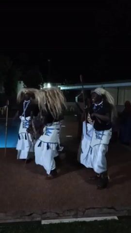 At night, five or six dark-skinned men dance on a dark brown tile or brick patio in front of a dark building with some windows, and, at the left, a lit blue swimming pool. The dancers wear black shirts, white, loose, patterned skirts, sandals, and long, straight, white hair. At least one carries a spear. The background is blurred.