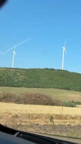 A video taken from inside a vehicle shows a scenic view of rolling hills under a clear blue sky. Two large white wind turbines stand prominently on the crest of a green, tree-covered hill. The turbine on the left has its blades in motion, creating a slight blur. A faint white contrail stretches diagonally across the sky above the left turbine, suggesting the presence of an aircraft. In the foreground, a field of golden wheat or barley stretches across the landscape, suggesting a rural setting and possibly a summer season. The lower part of the video shows the interior dashboard of the vehicle from which the footage is being recorded. The sun appears to be shining brightly, casting light on the scene. The image is slightly shaky, indicating it was likely filmed while the vehicle was in motion.