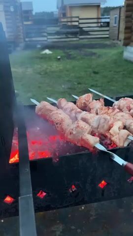 The video shows an outdoor cooking scene at dusk, likely in a rural or countryside setting with wooden log cabins visible in the background. The primary focus is a metal barbecue grill with glowing red coals at the bottom. Several metal skewers (shashlik) are placed over the coals, holding pieces of raw meat, likely pork or chicken, which are beginning to cook and show some browning. Smoke rises from the grill, indicating the cooking process. To the right of the skewers, a metal grill basket filled with other pieces of meat, possibly chicken wings or drumsticks, is also resting on the grill. A metal spatula with a red handle is visible, leaning against the grill. Towards the end of the video, a young boy wearing an orange vest and dark long-sleeved shirt comes into view, standing behind the grill, observing the cooking. The overall atmosphere suggests a casual and pleasant evening barbecue.