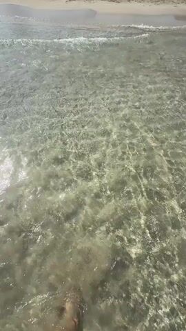 This media file shows a first-person perspective of someone walking in shallow, clear water. The sun is shining brightly, reflecting off the water's surface, creating shimmering patterns. The water appears to be at a beach, with soft sand visible beneath the surface. Gentle waves are rolling in, creating ripples and splashes around the person's feet. The person is wearing striped shorts. The overall scene conveys a sense of relaxation and enjoyment of a warm, sunny day at the beach, likely in Santa Eulària des Riu, Spain. The time of day appears to be daytime, with clear weather and a calm sea.