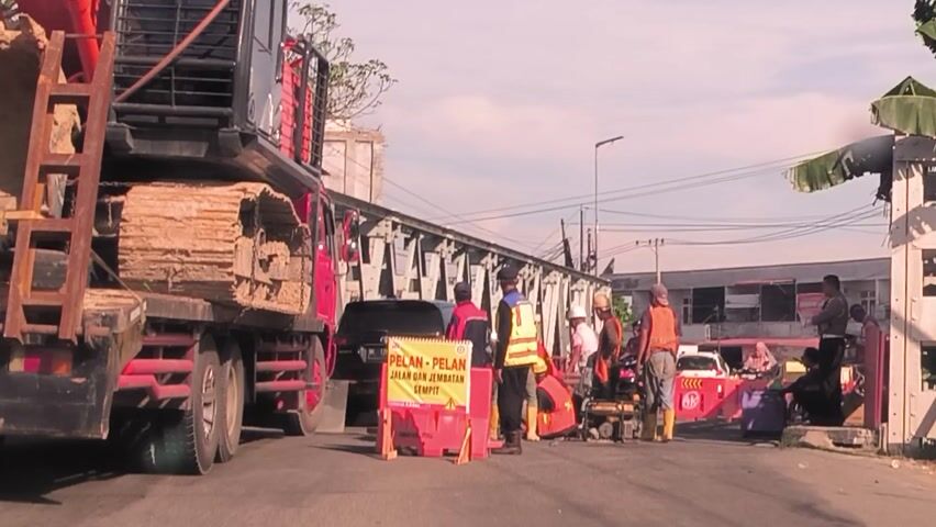 The video captures an outdoor scene in Kruet Teumpeun, Indonesia, depicting a construction or roadwork site. Several individuals, likely workers, are present; many are wearing high-visibility vests. A man in a yellow vest and blue hat is centrally positioned, seemingly overseeing or engaging in activity.

Two trucks are visible: a large, dark-colored truck with a flatbed carrying materials is on the right, and a smaller red truck is behind the workers. Attached to the smaller red truck is a prominent yellow banner with red text that reads "PELAN - PELAN JALAN DAN JEMBATAN SEMPIT TERIMA KASIH," which translates to "SLOW - SLOW NARROW ROAD AND BRIDGE THANK YOU." This sign suggests work is being done on a road or bridge with constricted passage.

Metal scaffolding or bridge support structures are visible on the left side of the frame. Overhead power lines or utility cables stretch across the clear, light blue sky, indicating an outdoor urban or semi-urban setting. The lighting suggests either early morning or late afternoon. Banana leaves are visible in the lower left corner. The individuals appear engaged in their tasks, with no strong emotions discernible.