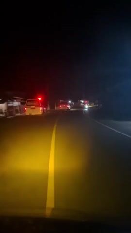 The video shows a dark street scene at night from the perspective of a vehicle with yellow headlights. A yellow line runs down the center of the road. Several vehicles are visible ahead, their taillights glowing red. One white vehicle, possibly an SUV or pickup truck, is directly in front, with its taillights on. Beyond it, more vehicles are seen with red taillights and some with flashing blue or white lights, possibly indicating emergency vehicles or a police presence, further down the road. Buildings are visible on the left side of the road, faintly illuminated by streetlights or other sources. The overall atmosphere is dark, with the bright yellow headlights of the recording vehicle providing the primary illumination for the foreground.
