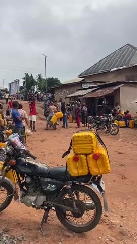 The video shows a large group of people gathered outdoors, most holding or carrying yellow jerry cans. The ground is red dirt. A motorcycle with four jerry cans strapped to the back sits in the foreground. A small water tower is visible in the background, surrounded by trees and buildings. People are milling around, some on motorcycles, most are dressed casually. The scene appears to be a distribution point, possibly for water or fuel. The sky is overcast.