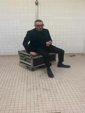 A man is sitting on a wheeled case, which appears to be a piece of travel luggage. He’s wearing glasses, a dark blazer, and dark trousers. He has a gray beard and a shaved head. He is holding a beverage in a clear plastic cup. The room is tiled in white and has a yellow border near the ceiling. The man looks at the camera and appears to speak with an exaggerated mouth movement.