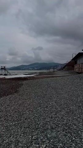 The scene depicts a gray, overcast day at a pebble beach. The foreground consists of a wide expanse of smooth, gray pebbles stretching towards the water's edge. In the mid-ground, waves are gently breaking against a concrete barrier and a wooden lifeguard tower with stairs leading up. Further back, across the water, a line of hills stretches across the horizon, partially obscured by clouds. To the right of the lifeguard tower is what appears to be a wooden structure. The sky is heavily clouded, adding to the overall somber mood of the scene. There are no people visible in the frame.