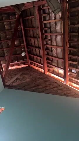 The video shows the underside of a roof structure. The roof is made of wooden beams and rafters, with some insulation material visible. Red clay tiles are used for roofing. A light bulb hangs from the ceiling. The walls below the roof appear to be painted a light blue-green color. The scene is indoors, possibly in a building under construction or renovation.