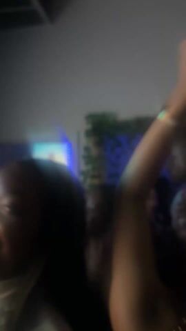 The video shows a crowded indoor space with people dancing and moving. In the foreground, a person with brown skin is wearing a light-colored shirt and smiling, looking upwards. To their right, another person is raising their arms, wearing a gold bracelet and a dark sleeveless top with a chain-like design. The background is blurry, showing hints of blue lighting, possibly a screen, and some greenery. The video is shaky and the focus is soft.