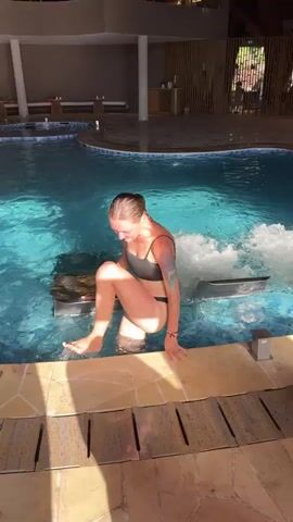A video shows a young woman exiting a swimming pool in what appears to be an indoor or covered resort setting, potentially in Mu Cang Chai, Vietnam. The woman, wearing a wet black bikini and sporting tattoos on her arms, is first seen squatting at the edge of the blue-tiled pool. She then stands up and steps out of the water, smiling cheerfully at the camera. As she walks closer, water drips from her body and bikini. She brings her hands to her chest, drawing attention to her wet black bikini top, while the camera zooms in on her torso. In the background, there are lounge chairs and a section of the pool with a water jet creating bubbles, suggesting a jacuzzi or massage area. The bright lighting indicates it is daytime. No visible text is present in the video.