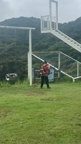 A person wearing an orange jacket and a backpack is standing on a grassy hill and appears to be cutting the grass with a weed whacker. There is a swing set to the left, a white staircase with a slide structure to the right, and lush green mountains in the background under a cloudy sky. Two cartoonish figures, possibly mascots, are standing in the grass further down the hill. The person is facing away from the camera and moving the weed whacker.