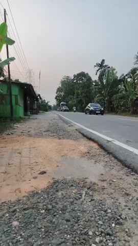 A roadside scene at dusk features a gray, overcast sky. The camera is positioned low to the ground, close to the edge of an asphalt road. The road appears to be in fair condition, with a slightly uneven surface and a worn curb. To the left of the road, there is a dirt and gravel shoulder with some patchy vegetation. Further left, a simple, small building with green siding and a corrugated metal roof is visible. In the background, lush green trees and palm trees line the right side of the road. Several vehicles are traveling on the road. A black SUV is in the foreground, moving to the right. Further down the road, a blue and green truck is traveling in the same direction. A person on a motorcycle is also visible further in the distance. Utility poles and power lines are present on the left side of the frame. The overall impression is of a rural or semi-rural road at the end of the day.