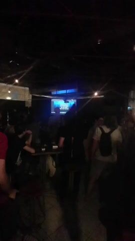 The video captures a dimly lit room, possibly a bar or karaoke venue. In the center, a large screen displays lyrics with a blue background. Several people are present, mostly blurred or in silhouette due to the low light. A projector is visible hanging from the ceiling, aimed at the screen. Some individuals are gathered around tables, while others stand and appear to be watching the screen or interacting with each other. The atmosphere seems casual and social, with hints of entertainment.