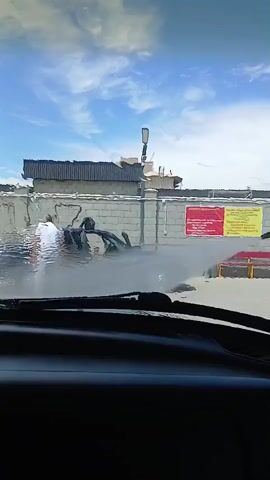 A man is power washing a car. The car's doors are open, and the interior appears to be damaged. The man is holding a power washer and spraying water on the car. There is another person standing near the car, partially obscured by the water spray. The scene takes place outdoors in front of a building with a brick wall. The sky is blue with some clouds.
