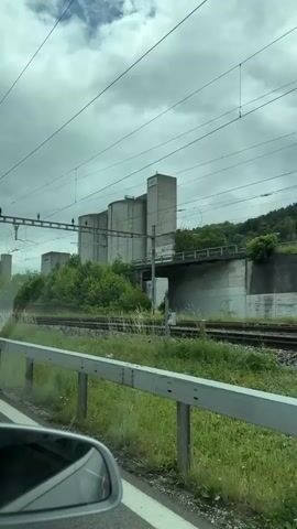 The video captures a moving shot from inside a vehicle, revealing an industrial landscape near Zurzach, Switzerland, on an overcast day. In the foreground, a metal barrier separates the road from green grass and a set of train tracks. Beyond the tracks, large, grey, cylindrical and rectangular concrete buildings, resembling silos or industrial structures, dominate the mid-ground. One building clearly displays the text "L.G.Z." Overhead power lines, likely for an electric train system, crisscross the cloudy sky. Further in the background, amidst lush green trees and bushes, several residential houses with dark roofs are visible on a rising incline. A car's rearview mirror is partially visible in the bottom left corner of the frame. The scene suggests a transition from a road to a railway and industrial area, with residential zones nearby.